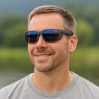 Man wearing Deep Gray polarized sunglasses with reflective blue-tinted lenses, standing outdoors near a lake. He’s smiling casually, dressed in a light gray t-shirt, with a calm natural landscape in the background.