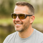 Man wearing Lime Shift polarized sunglasses with amber-yellow tinted lenses, smiling outdoors in a natural setting. He is dressed in a light gray t-shirt with soft, blurred greenery in the background.